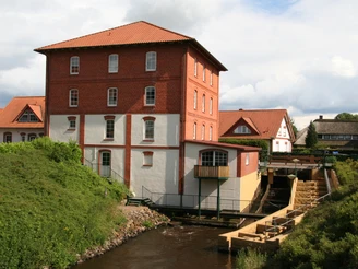 Wassermühle Liebenau Wassermühle mit rotem Ziegelbau und Flusslauf, umgeben von grüner Landschaft und bewölktem Himmel.