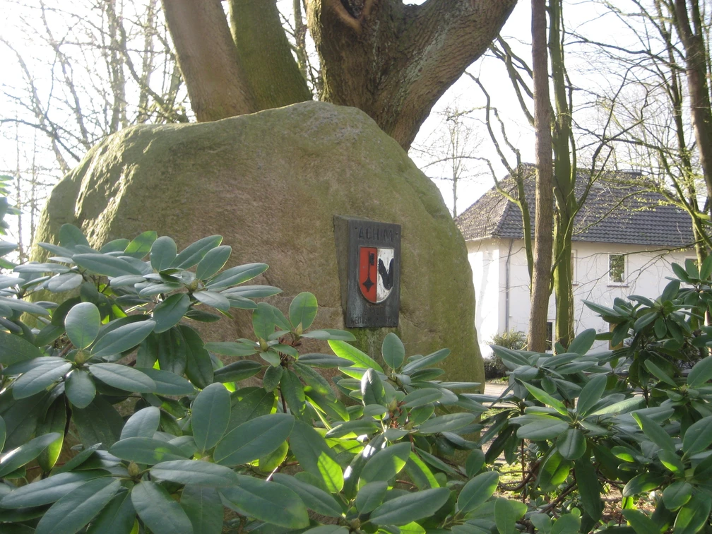 Großer Findling mit Wappen im grünen Rathausgarten, umgeben von Rhododendron und Bäumen.