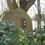 Großer Findling mit Wappen im grünen Rathausgarten, umgeben von Rhododendron und Bäumen.