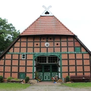 Backsteinfachwerkhaus mit rotem Ziegeldach und grüner Holzverzierung vor Wiese und Bäumen.