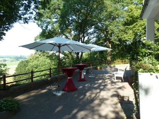 Haus Hünenburg Terrasse mit Sonnenschirmen, Stehtischen in Rot und Blick auf grüne Landschaft unter Bäumen.