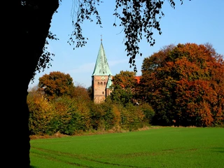 Felicianus Kirche Weyhe hinter buntem Herbstlaub, Spitzdach mit Turmspitze und Wiesen im Vordergrund.