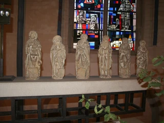 Sechs steinerne Apostelfiguren stehen in einer Kirchenreihe vor farbigem Glasfenster in Nienburg.