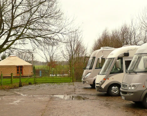 Wohnmobile stehen nebeneinander auf einem befestigten Stellplatz am Rand eines grünen Feldes.