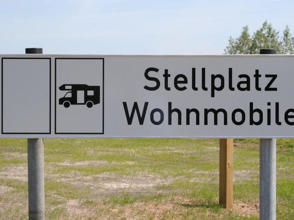 Wohnmobilstellplatz Schild für Wohnmobilstellplatz auf grünem Rasen vor blauem Himmel mit Bäumen im Hintergrund.