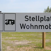 Wohnmobilstellplatz Schild für Wohnmobilstellplatz auf grünem Rasen vor blauem Himmel mit Bäumen im Hintergrund.