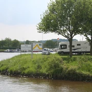Wohnmobilstellplatz Marina Mehlbergen Wohnmobil auf begrüntem Platz an einer Marina, mit Bäumen und ruhigem Wasser im Hintergrund.