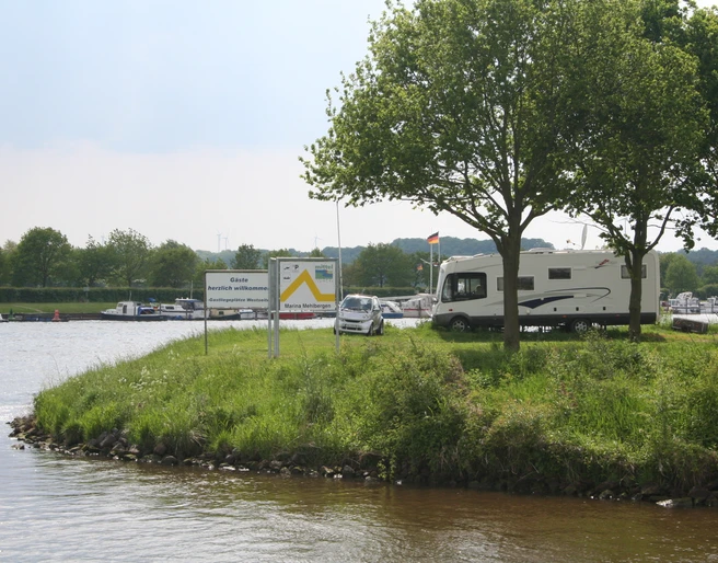 Wohnmobilstellplatz Marina Mehlbergen Wohnmobil auf begrüntem Platz an einer Marina, mit Bäumen und ruhigem Wasser im Hintergrund.