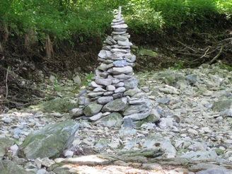 Eine Pyramide aus gestapelten Steinen steht im trockenen Flussbett, umgeben von dichtem Wald.