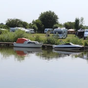 Wohnmobile und Boote am idyllischen Flussufer, umgeben von Bäumen und grünen Wiesen.