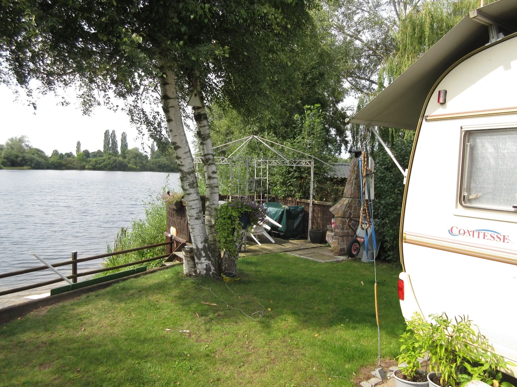 Stedeberger See Grüne Wiese mit Wohnwagen am Seeufer, umgeben von Bäumen und einem Pavillon im Hintergrund.