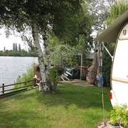 Stedeberger See Grüne Wiese mit Wohnwagen am Seeufer, umgeben von Bäumen und einem Pavillon im Hintergrund.