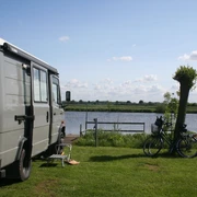 Wohnmobil steht auf einer Wiese am See, neben zwei Fahrrädern und einem Stuhl mit Blick aufs Wasser.