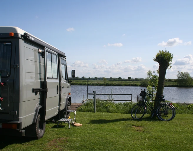 Wohnmobil steht auf einer Wiese am See, neben zwei Fahrrädern und einem Stuhl mit Blick aufs Wasser.