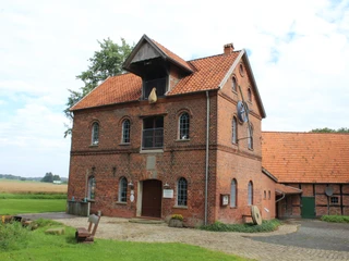 Wassermühle Süstedt Rote Backstein-Wassermühle mit Satteldach, umgeben von Grünflächen und einem gepflasterten Weg.