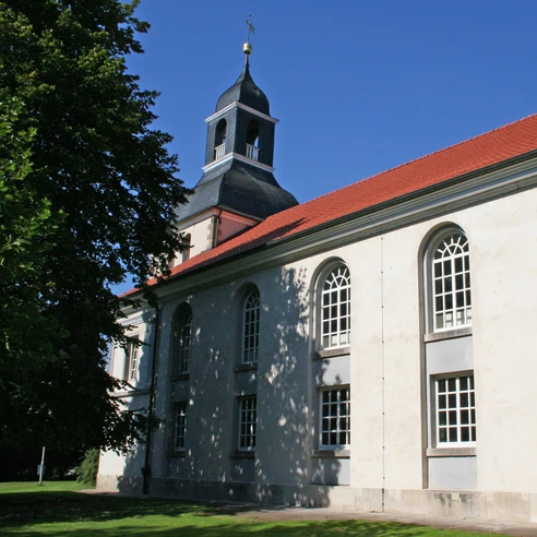Kirche Blender Eine Kirche mit rotem Ziegeldach und weißer Fassade, umgeben von Bäumen und blauem Himmel.