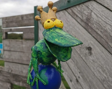 Froschkönig am Lieblingsort Holtgast Eine bunte, humorvolle Froschskulptur mit Krone auf einem blauen Ball vor einem Holzzaun.