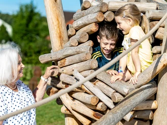 Ludowinengarten in Bökendorf Kinder spielen in einem hölzernen Klettergerüst, während eine ältere Frau sie lächelnd beobachtet.