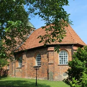 Rundziegelbau der Kirche Binnen mit rotem Dach vor einem strahlend blauen Himmel und umgeben von Bäumen.
