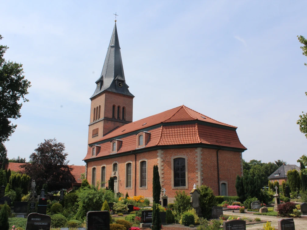 Kirche Schwarme Backsteinkirche mit spitzem Turm vor einem blauen Himmel, umgeben von einem grünen Friedhof.