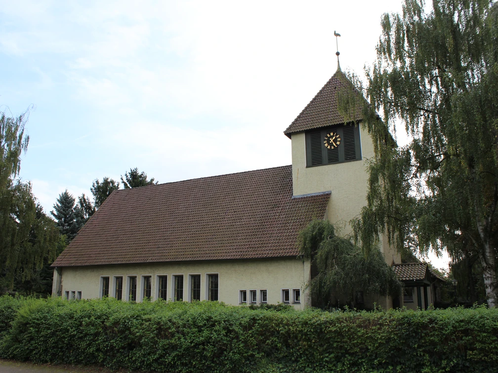 Kirche in Essern mit markantem Glockenturm, umgeben von Bäumen und von einer Hecke eingefasst.