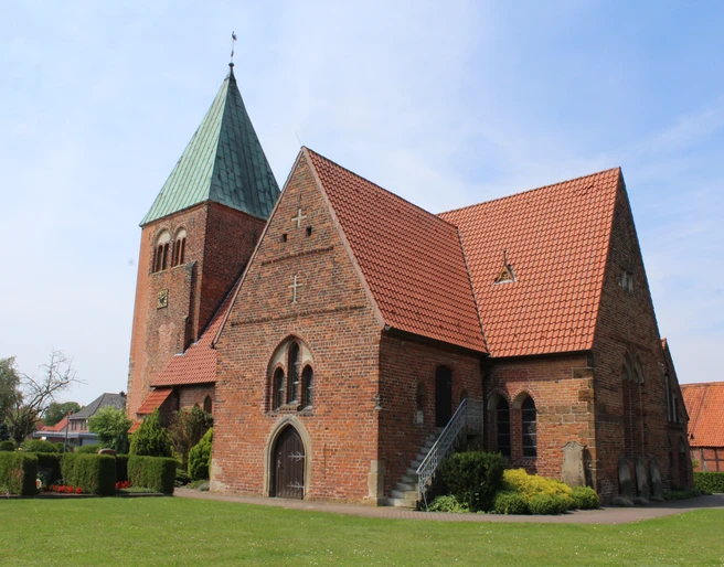 Backsteinkirche mit grünem Turmdach, Satteldächern und gepflegtem Grün vor blauem Himmel.