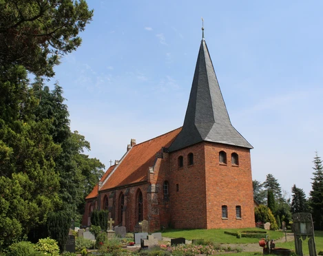 Backsteinkirche St. Crucis in Hoyerhagen mit spitzem Kirchturm umgeben von Bäumen und einem Friedhof.