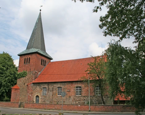 Kirche Holtorf Backsteinkirche mit markantem Spitzdach, umgeben von Bäumen, vor bewölktem Himmel in Holtorf.