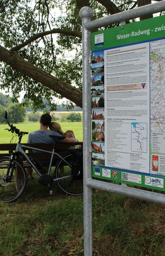 Radfahrer am Weser-Radweg Zwei Radfahrer pausieren an einer Bank auf einer grünen Wiese, neben einem Info-Schild am Weser-Radweg.