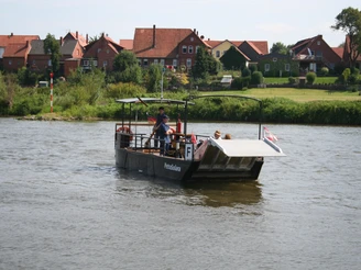 Fähre Petra Solara Windheim-Hävern Fähre überquert Fluss mit Häusern im Hintergrund, Passagiere und der Fährmann an Bord.