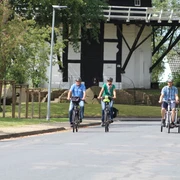 Drei Radfahrer in sportlicher Kleidung fahren eine asphaltierte Straße entlang, im Hintergrund die Achimer Windmühle.
