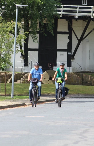 Radfahrer an der Achimer Windmühle Drei Radfahrer in sportlicher Kleidung fahren eine asphaltierte Straße entlang, im Hintergrund die Achimer Windmühle.