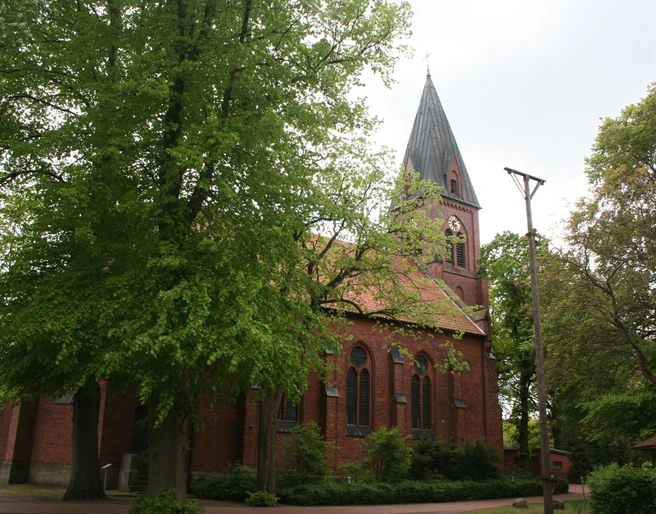 St. Vitus-Kirche Schinna, eine Backsteinkirche mit hohem Turm, umgeben von Bäumen, Himmel bedeckt.