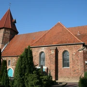 St. Laurentius-Kirche Backsteinkirche mit rotem Satteldach, spitzem Turm und grüner Holztür, umgeben von Bäumen und Pflanzen.