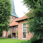 Backsteinkirche mit grünem Spitzdach, flankiert von großen Laubbäumen, auf einer gepflegten Rasenfläche.