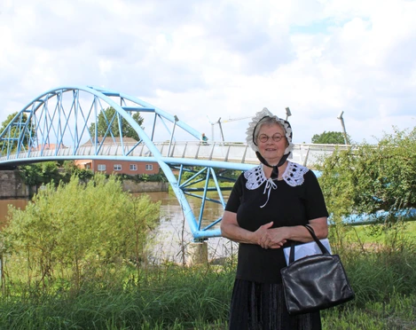 Gewandete Stadtführerin Nienburg Eine Frau in historischer Tracht steht vor einer blauen Brücke an einem Flussufer in Nienburg.