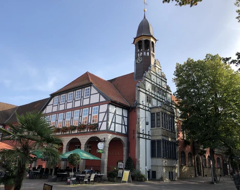 Rathaus Nienburg mit Fachwerk, rotem Backstein, Uhrturm, umgeben von Bäumen und Sonnenschirmen.