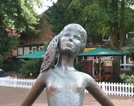 Bronzestatue eines Mädchens mit Zöpfen im Freien, umgeben von Bäumen und einem Café im Hintergrund.