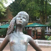 Kleine Nienburgerin  Bronzestatue eines Mädchens mit Zöpfen im Freien, umgeben von Bäumen und einem Café im Hintergrund.
