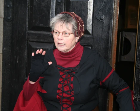Ältere Frau in mittelalterlicher Kleidung steht vor einer Holztür und hält die Hand erhoben.