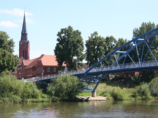 Backsteinbauten mit hoher Kirchturmspitze hinter einer blauen Brücke über einem ruhigen Fluss.