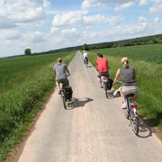 Radfahrer auf einem schmalen Weg entlang grüner Felder, blauer Himmel mit weißen Wolken oben.