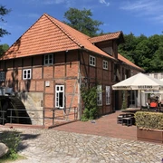Klostermühle Heiligenberg Historische Mühle mit rotem Fachwerk, umgeben von Bäumen und Sonnenschirmen im Außengastronomiebereich.