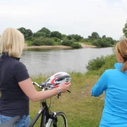 Zwei Frauen stehen mit Fahrrädern am Weserufer, blicken auf den ruhigen Fluss und die Natur.