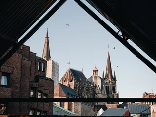 Aachener Dom Der Aachener Dom ragt in den Himmel, eingefasst in ein Gerüst aus alten Gebäuden und modernen Strukturen. Seine gotischen Türme thronen über der Umgebung und fangen das Licht des klaren Himmels.