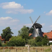 Historische Windmühle, umgeben von grüner Vegetation und roten Ziegeldächern unter blauem Himmel.