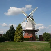 Windmühle unter blauem Himmel mit weißen Wolken, umgeben von grünen Bäumen und Wiesen.