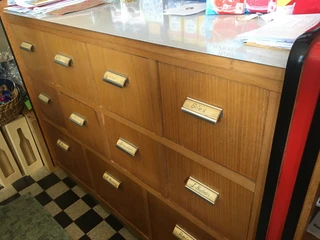 Tante Emma Neuenkruge Auslage.jpg Das Bild zeigt eine Schubladenwand mit Etiketten in einem Tante-Emma-Laden. Die Schubladen aus Holz sind in einem klassischen, rustikalen Stil gehalten und jeweils mit einem kleinen Metallschild beschriftet. Einige der Beschriftungen sind "Grieß", "Sabine", "Oma".```htmlHolzschubladen mit Beschriftungen, Teil eines Ladens.