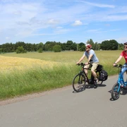 Radfahrer Zwei Radfahrer fahren bei sonnigem Wetter auf einer asphaltierten Straße durch eine grüne Landschaft.