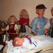 Puppenmuseum Eine Gruppe fröhlicher Puppen in traditioneller Kleidung umgibt einen Kinderwagen mit einem Baby.